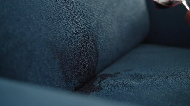 Close-up male hands dropping glass of red wine on blue couch, spilling wine and creating stain, depicting accident at home, party, celebration. Spilling wine from glass onto couch creating large stain