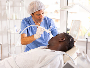 Experienced male aesthetician applying atomized nutrients with airbrush onto black man face to nourish and rejuvenate skin. Modern hardware cosmetology