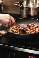 Hand of a person stirring sliced sausage in a pan while cooking on the stove, with a visible pot in the background