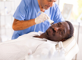 Fototapeta premium Adult African American undergoing high frequency ultrasound therapy to reduce fine wrinkles and rejuvenate facial skin by stimulating collagen production. Modern cosmetology and male grooming concept
