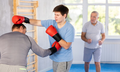 Determined guy mastering self-defense techniques at boxing gym, practicing punches in sparring with...
