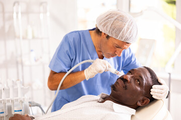 Fototapeta premium Adult African American undergoing facial treatment using ion bubble attachment for gentle exfoliation, deep hydration and skin revitalization at cosmetic medicine clinic. Male grooming concept