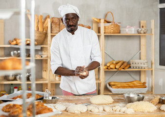 Skilled black baker in white uniform carefully handling individual dough portions at flour-covered...