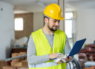 An architect in protective hard hat and yellow vest checks room renovation plan using a laptop