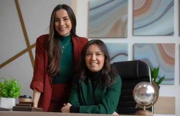 Obraz premium Two professional businesswomen smiling confidently in a modern office, one sitting at a desk and the other standing beside her, symbolizing leadership, teamwork, and success.