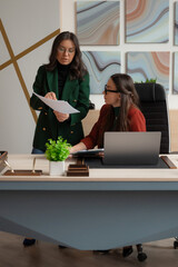 Two businesswomen in a modern office discussing documents, one standing and explaining, the other...