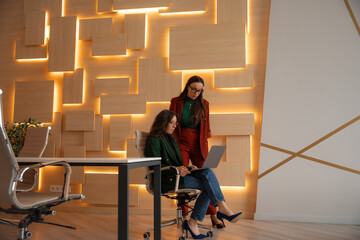 Two businesswomen working in a modern office, one sitting with a laptop, the other standing and...