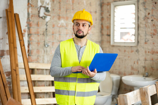 Focused young bearded architect in yellow protective workwear using modern software to create rooms design while standing in house under reconstruction