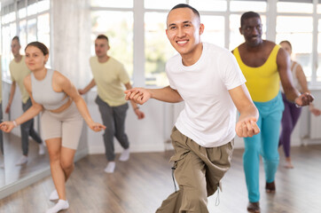 Sporty smiling young Asian guy practicing rhythmic aerobics during group workout in spacious...