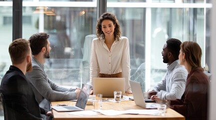 A smiling young businesswoman leads a diverse team meeting around a large table in a modern office, using laptops and documents