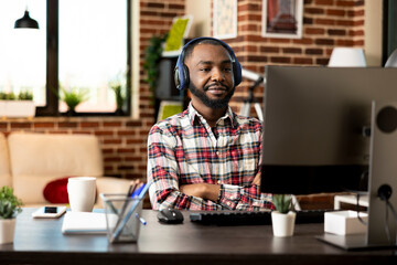 Black male freelancer seated with headphones and arms crossed, watching online webinar on desktop...