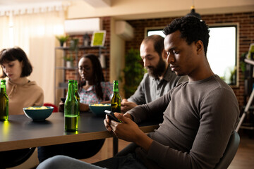 Selective focus on african american man holding smartphone, with bottle of beer on table beside him. Young male adult browses online while sitting with diverse friends in cozy room.