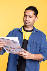 Indian young guy browsing though financial audit report paperwork, feeling overworked and confused...