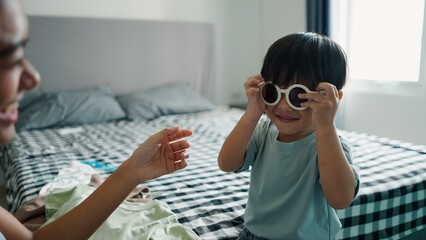 Cheerful kid wearing oversized sunglasses, playing near mother preparing travel wardrobe on bedroom bed