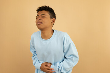 Brown skinned Latino teenager touching his stomach with his hands, with an expression of stomach pain on a light brown background with copy space, young man with abdominal pain
