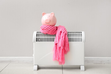 Piggy bank with warm scarf on electric convector heater near white wall at home. Heating saving...
