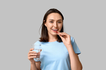 Pretty young woman with pill and glass of water on grey background
