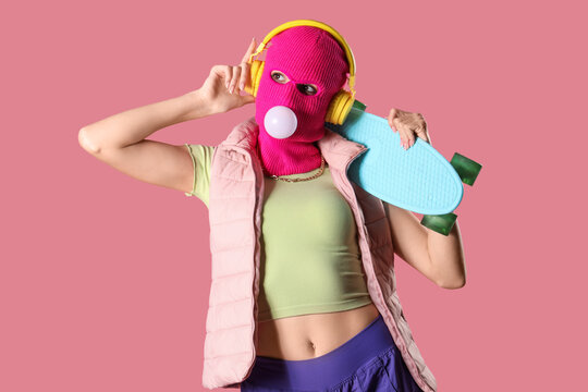 Beautiful young stylish woman in balaclava with headphones and skateboard chewing gum on pink background