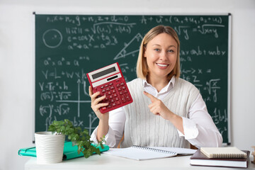 Mature female math teacher pointing at calculator and sitting at table with notebooks in classroom