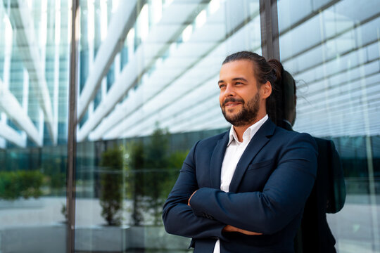 Confidence successful businessman in suit with beard standing in front of office glass building lean on wall arm crossed looking away and smile. Hispanic modern business man portrait. Business suit