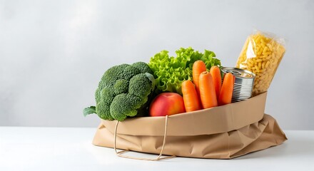 A brown paper bag filled with fresh produce: broccoli, lettuce, carrots, apple, and packaged pasta and canned food. On a white surface against a light background.