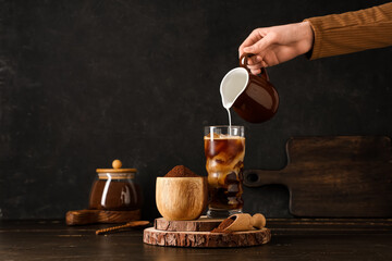 Woman adding milk to iced coffee on table with bowl and scoop with powder