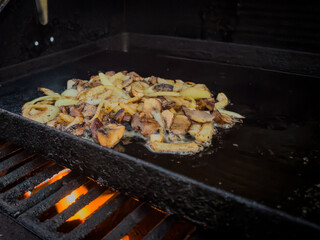 A black cast iron frying pan with chopped yellow onions frying over an open flame on a barbecue. The caramelized vegetable is a topping for various barbecue meats. The grilling is with soft butter.