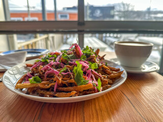 A large white plate filled with shoestring French fries deep fried and crispy covered with cheese sauce, pickled onions and cilantro. There’s a white porcelain cup and saucer with coffee on the table.