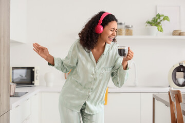 Morning of young African-American woman with headphones and cup of coffee singing in kitchen