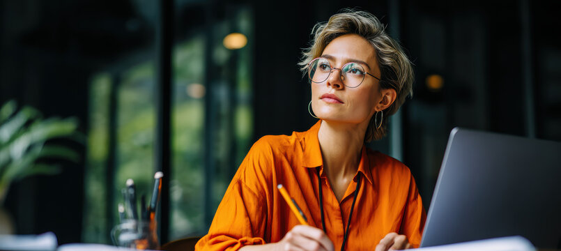 Thoughtful woman in orange shirt works at desk with laptop and pencil, surrounded by creative workspace