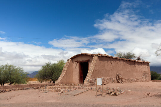 Main adobe chapel with sloping walls and a cartwheel, at El Mayorazgo Complex, Tinogasta, Catamarca. An icon of the Adobe Route under a blue sky with clouds.