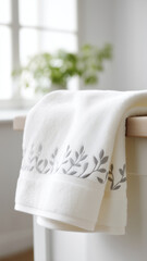 Soft white towel with gray leaf embroidery draped over wooden surface in bright room with blurred green plant background