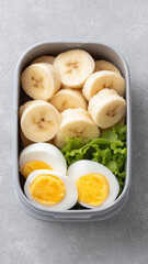 Sliced banana and boiled egg with fresh green lettuce in gray container, healthy and nutritious meal option for breakfast or snack