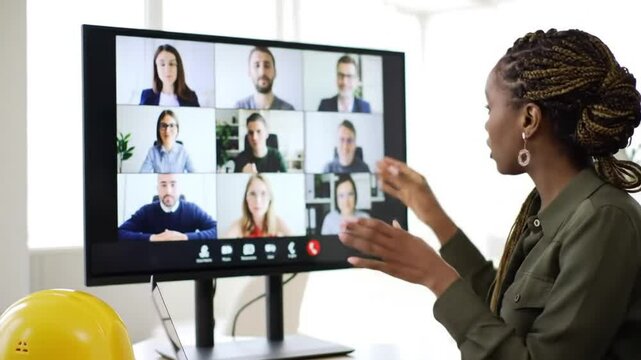 Black Woman Presents on Video Conference with Multiple Participants on Screen in Bright Office with Yellow Helmet