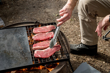 Man grilling beef steaks on a cast iron grill plate on a camp fire. Campfire cooking. Outdoor BBQ