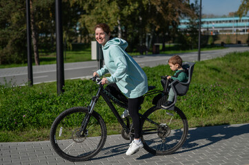 Caucasian woman riding bicycle with toddler son sitting behind her in child seat in park. 