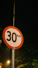 The thirty kilometers per hour speed limit sign stands illuminated against dark background at night.