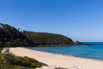 An idyllic sandy beach with aqua turquoise ocean water alongside a tree covered headland, captured on the South Coast of New South Wales, Australia.
