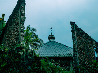 A mosque dome is partially visible behind old, ruined brick walls under a cloudy sky.