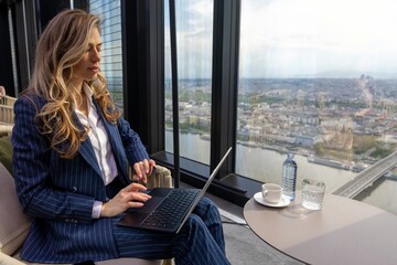 A dedicated businesswoman, dressed in a sleek and stylish suit, diligently works on her laptop in a highrise building, enjoying a breathtaking view of a vibrant and bustling city skyline
