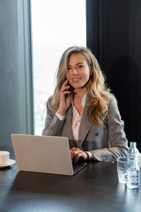 In a modern office, a confident businesswoman talks on her smartphone while working on her laptop,...
