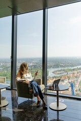 A woman sits comfortably and relaxed in a stylish, modern office space, engaging with her smartphone while enjoying a breathtaking city view filled with stunning skyscrapers and clouds