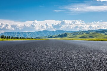 Fototapeta premium Wide asphalt road crossing a green valley with a mountain range and blue sky with clouds in the background