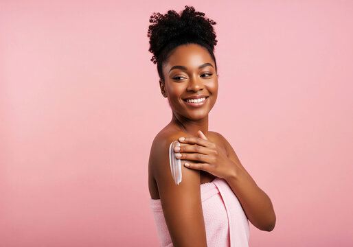A young African American woman smiles while applying body lotion to her shoulder. Wrapped in a pink towel, she stands against a soft pink background, radiating confidence and healthy skin.


