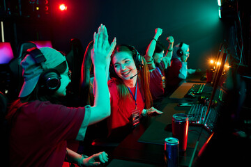 Group of multiethnic teenage girls wearing headsets sitting at gaming computers celebrating victory...