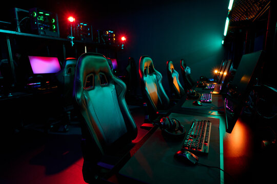 Empty gaming chairs and computer setups arranged in esports arena, illuminated by colorful lights, showing professional gaming environment prepared for girls gamers team competition