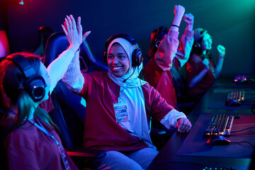 Diverse team of teenage girls sitting at gaming computers celebrating victory, wearing headsets and team shirts, smiling and giving high fives during esports competition