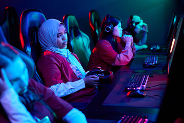 Diverse team of teenage girls sitting at gaming computers participating in e-sports competition,...