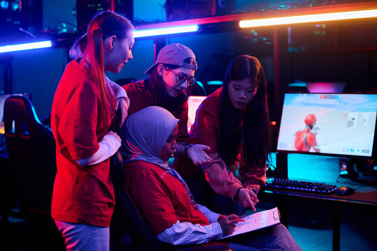 Group of diverse teenage girls collaborating during esports gaming session, standing and sitting around computer monitors, discussing strategy and analyzing gameplay on digital tablet