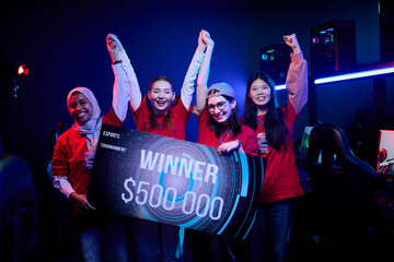 Diverse group of teenage girls celebrating esports tournament victory, holding large winner sign with prize amount, standing together in gaming room, smiling and raising arms in excitement © pressmaster